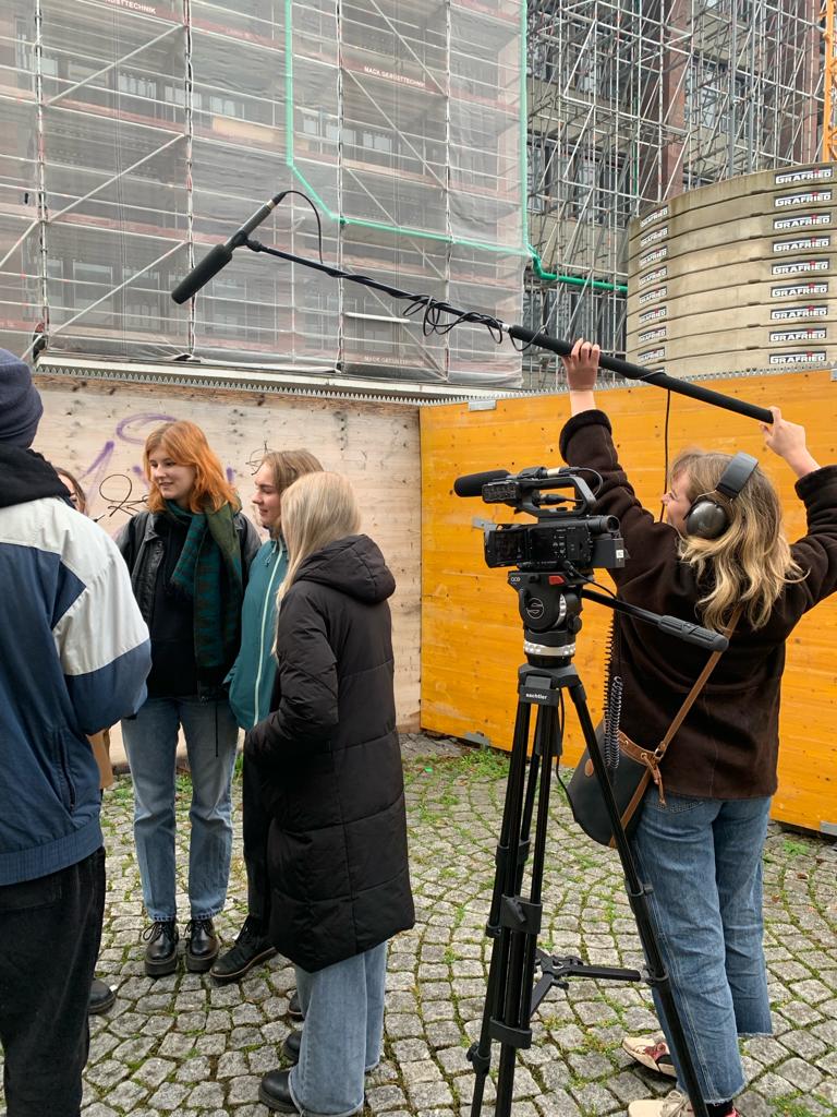 Kollegiengebäude 4, vor dem Café Senkrecht. Die Tonfrau hängt die Tonangel über die Schauspieler*innen. Die Kamera steht auf einem Stativ. Im Hintergrund ist eine Baustelle zu sehen.