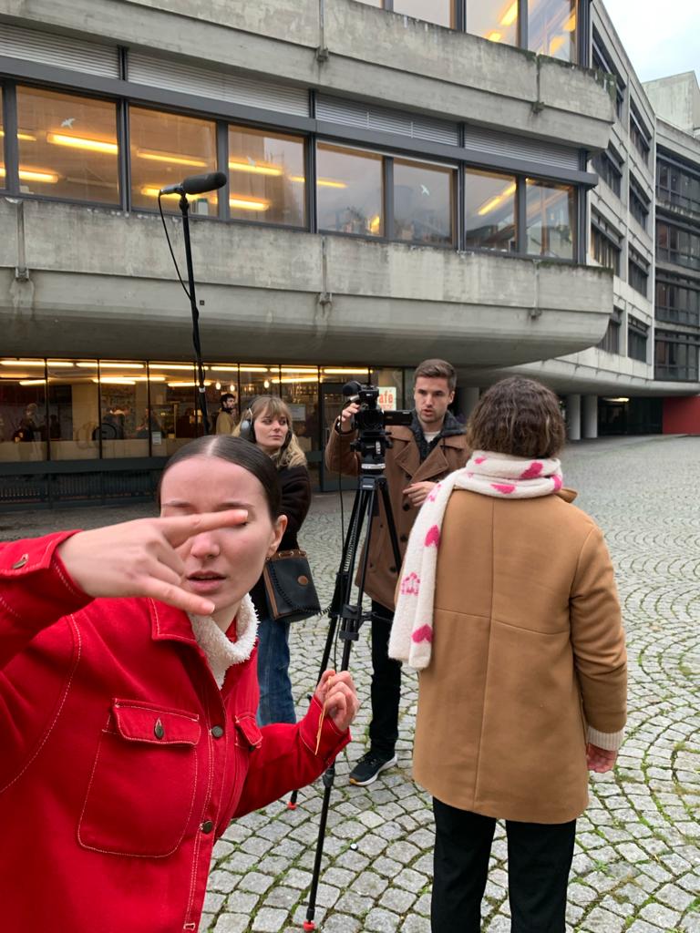 Vor dem Cafe Senkrecht beim Kollegiengebäude 4. Die Filmcrew steht mit Kamera und Tonangel bereit. Die Protagonistin zeigt eine Rock n Roll Handgeste in die Kamera