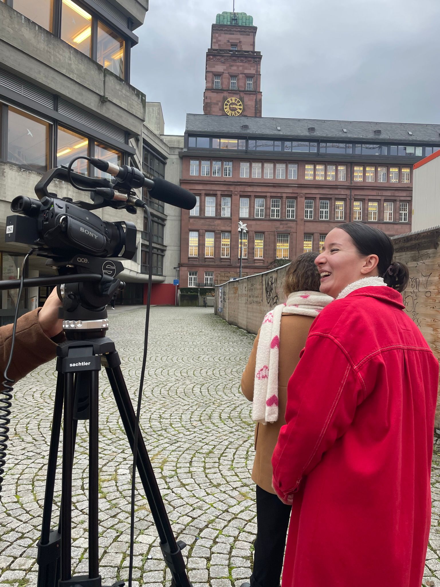 Zu sehen ist die aufgebaute Kamera und die Protagonistin des Films beim Café Senkrecht am KG 4 der Universität Freiburg. Die Protagonistin lacht.