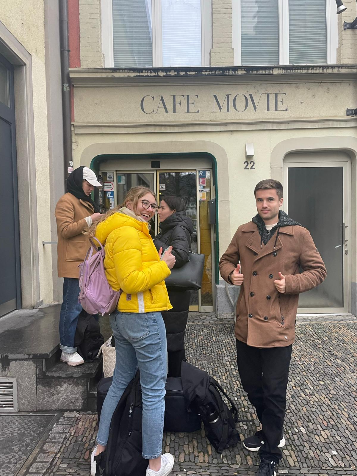 Die Filmcrew steht vor dem Café Movie in Freiburg und zeigt Daumen hoch in die Kamera. 