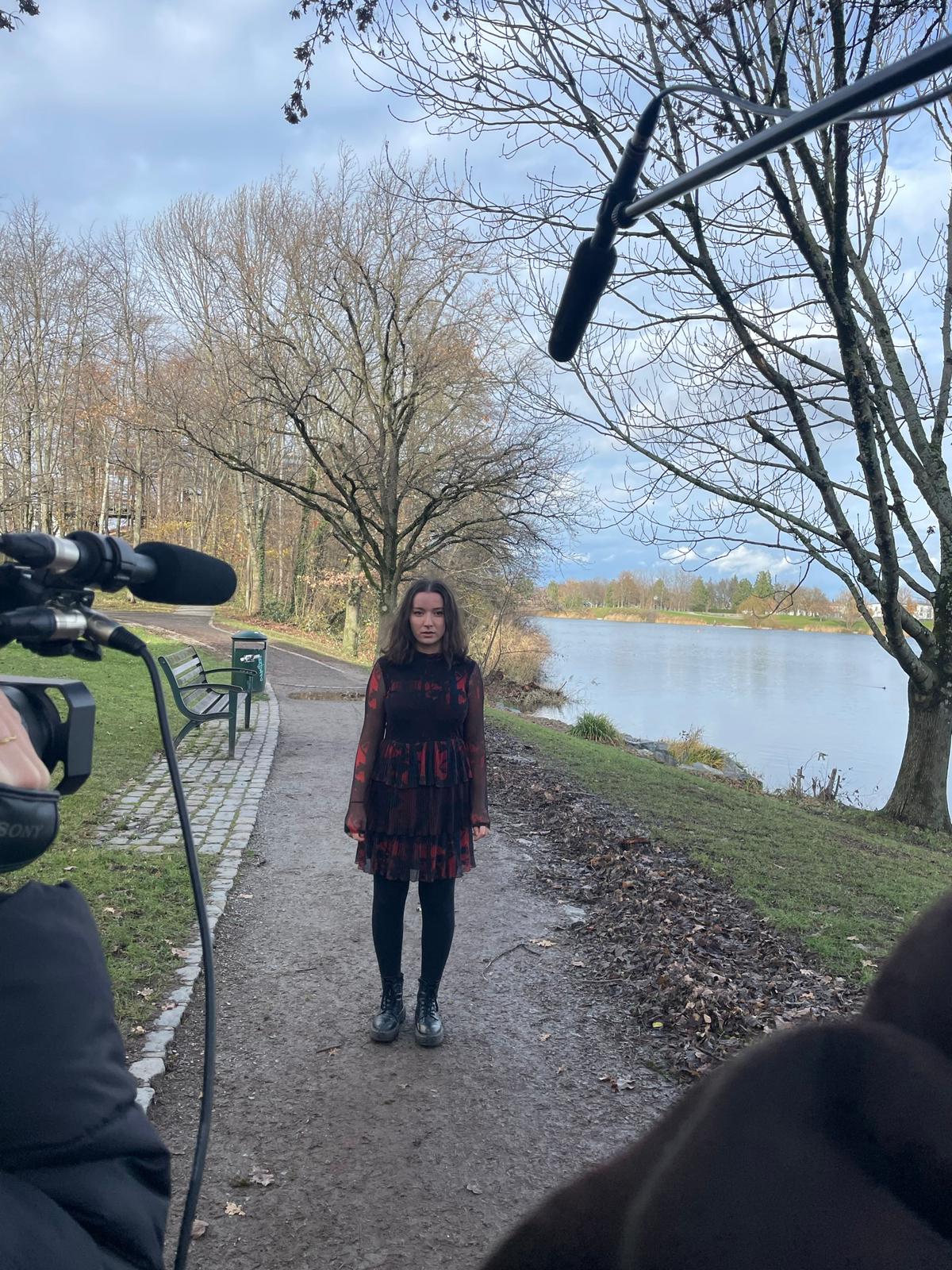 Zu sehen ist die Protagonistin auf einem Spazierweg am Seepark. Sie schaut Richtung Kamera. Die Kamera und die Tonangel ragen ins Bild.