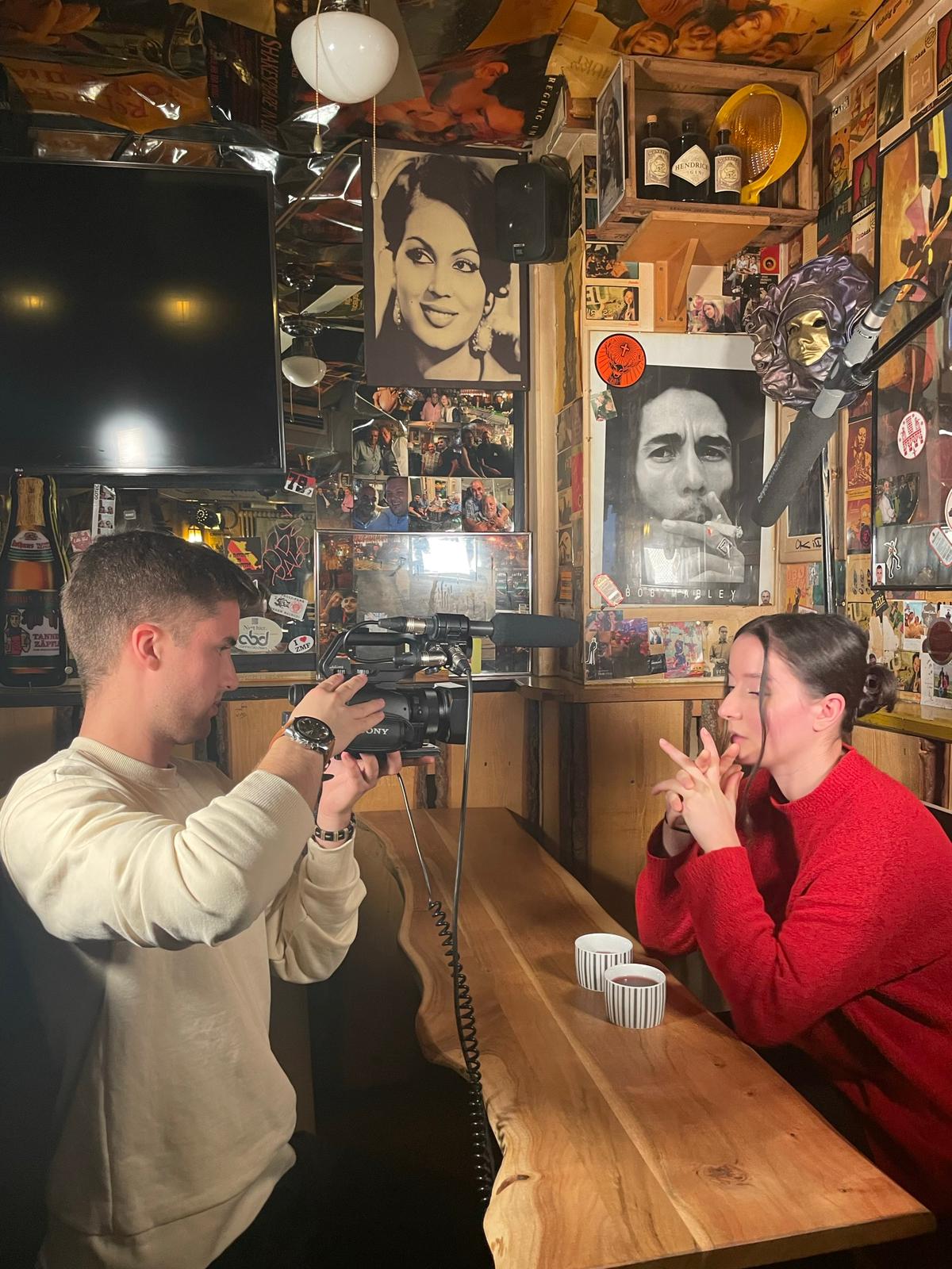 Making-Of Bild zeigt Kameramann, der die Protagonistin des Films filmt. Sie sitzen an einem Tisch in einer Bar. Die Wand ist mit mehren Postern von populären Musiker*innen geschmückt. Das Licht ist hell. Der Tisch ist aus Holz. Die Wand ist außerdem mit Holz verkleidet.